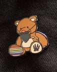 Enamel pin of a bear with a rainbow ball on a black denim background