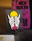 Pin with hands covering a face and a pink cross on a dark background with dice and a book.