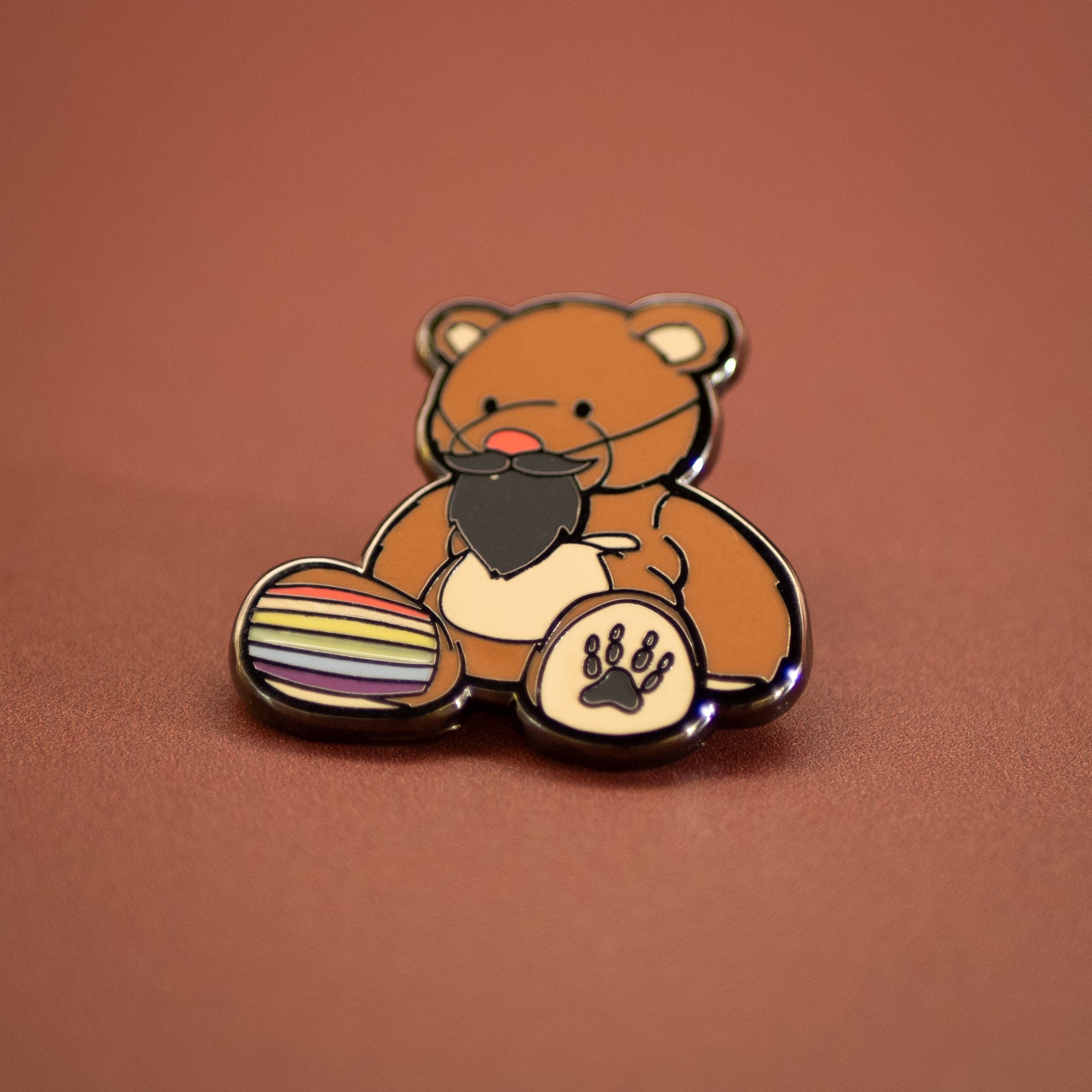 Enamel pin of a bear holding a rainbow flag and paw print on a brown background