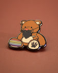Enamel pin of a bear holding a rainbow flag and paw print on a brown background
