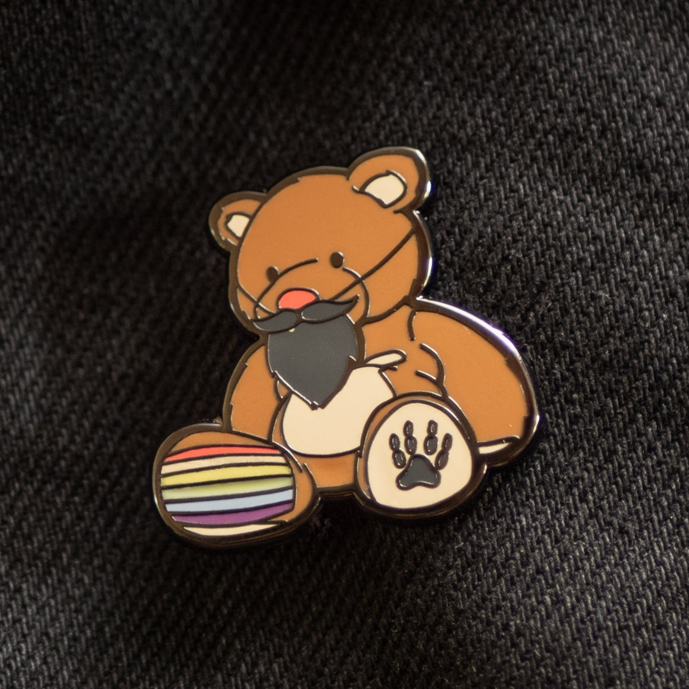 Enamel pin of a bear with a rainbow ball on a black denim background