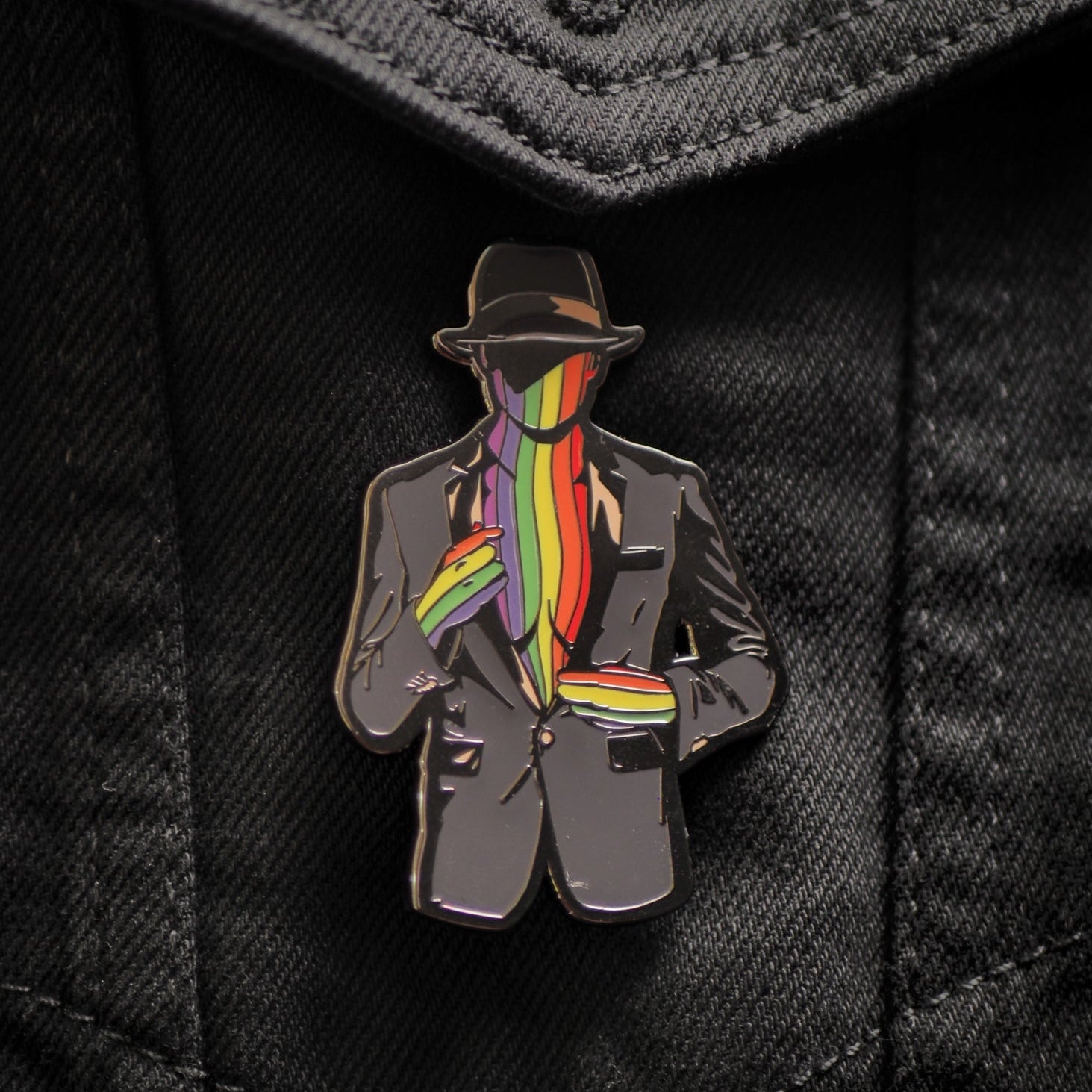 Pin with a design of a person wearing a suit and hat with a rainbow scarf on a black denim background