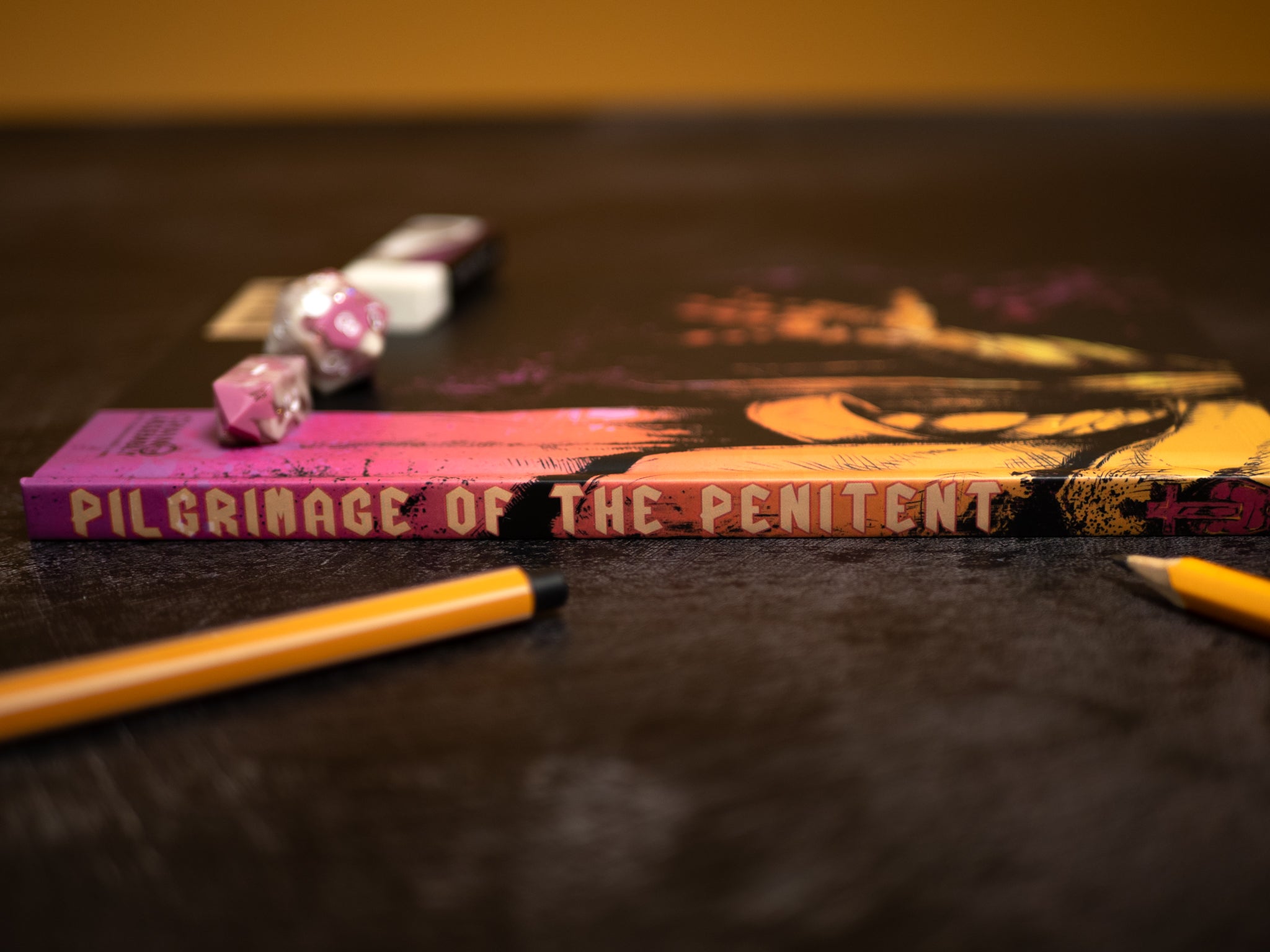 Book titled 'Pilgrimage of the Penitent' on a dark surface with pencils and decorative items.