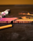 Book titled 'Pilgrimage of the Penitent' on a dark surface with pencils and decorative items.