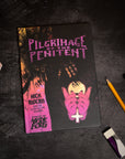 Book titled 'Pilgrimage of the Penitent' on a dark surface with pencils and crystals.