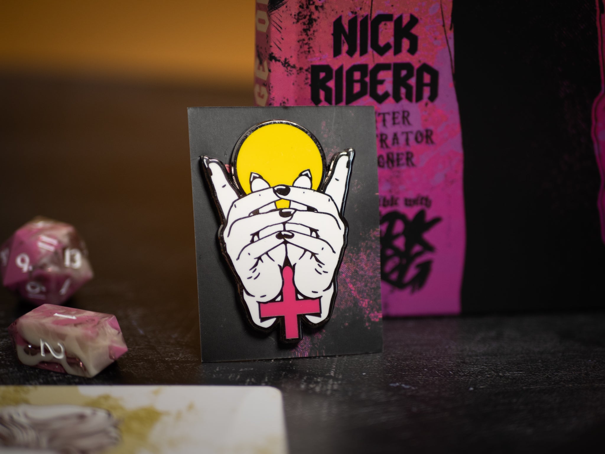 Pin with hands covering a face and a pink cross on a dark background with dice and a book.